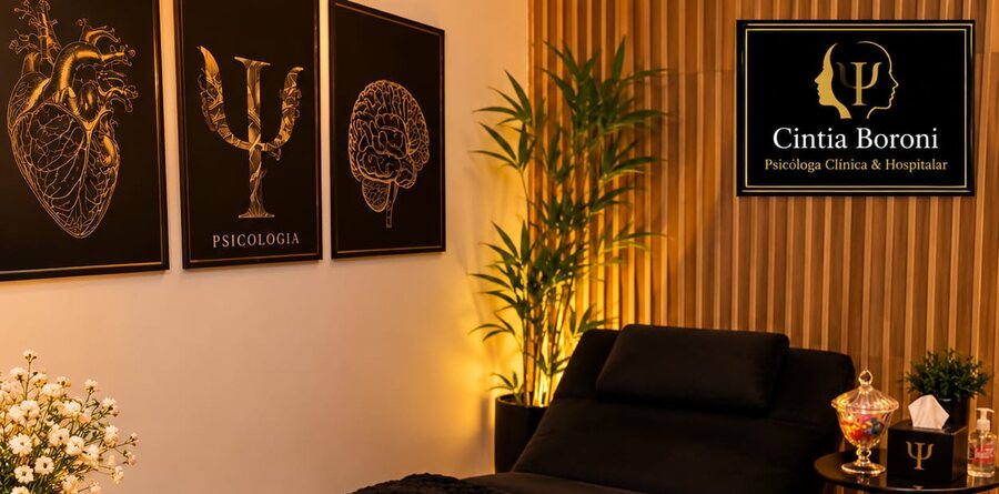 Sala de acolhimento do consultório, com divã escuro, quadros dourados e a placa da marca Cintia Boroni.